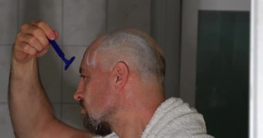 Banyoda bir adam tıraş bıçağı kullanarak kel kafasını tıraş ediyor. Bakım rutinine odaklanmış durumda ve güne hazırlanırken temiz bir görünüm sağlıyor..