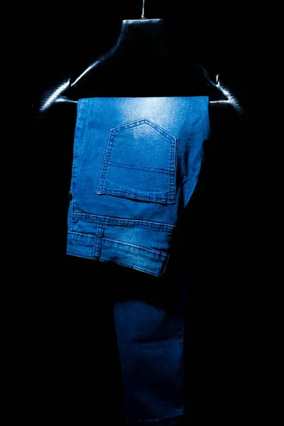 Kot pantolonun dinamik bir fotoğrafı, siyah arka plandaki askıda sergileniyor. Moda siteleri, reklamcılık ve editoryal kullanım için ideal.