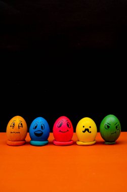 El çizimi emoji ifadeleri içeren beş canlı Paskalya yumurtası, dramatik siyah bir arkaplanı olan turuncu bir zemine yerleştirilmiş. Mizah, yaratıcılık ve Paskalya kutlaması için ideal.