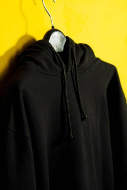 Siyah bir kapüşonlunun, temiz sarı bir arkaplanda asılı durduğu, modaya uygun ve canlı bir görsel cazibe yaratan minimalist bir fotoğraf..