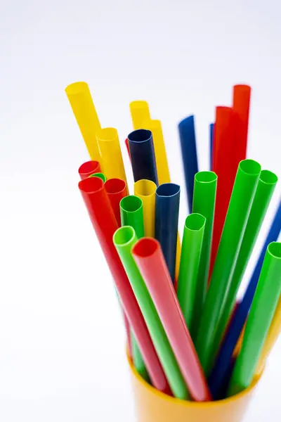 Bir grup renkli plastik pipet, düz beyaz arka plana karşı sarı bir fincanda dikey olarak dizilmiş..