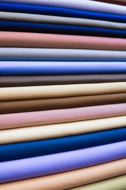 Yakın plan bir fotoğrafta çok sayıda kumaş rulosu düzgünce istiflenmiş, çeşitli renkler ve ince doku varyasyonlarını ortaya koyuyor. Yuvarlaklar sıkıca dizilmiş, yatay çizgide görsel olarak çekici bir şekil yaratıyor.