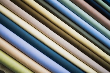 Yakın çekimde, köşegen çizgilerde sıralanmış sayısız kumaş rulo, sessiz toprak tonları ve serin mavi renkler sergiliyor. Ekmekler sıkıca sarılmış, kumaşların ince dokusunu ve parıltısını gösteriyor.. 