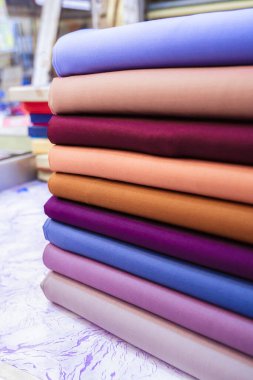 Düzgünce istiflenmiş çeşitli renkli kumaşların yakın plan görüntüsü. Ön planda, mor, leylak, açık mavi, şeftali ve kahverengi de dahil olmak üzere bir dizi pastel ve zengin renkte on adet kumaş var. Arka plan biraz bulanık.