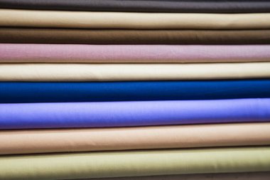 Yakın çekim titizlikle düzenlenmiş kumaş rulo yığınını ortaya çıkarır, çeşitli renk ve dokulardan oluşan bir palet sergiler. Her rulo eşsiz bir renk sergiler, yumuşak pastellerden zengin, derin tonlara kadar, çeşitliliği vurgular.