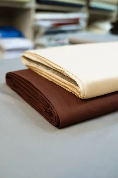 Gri bir yüzeyde düzgünce sarılmış iki parça kumaş. Üst rulo kremalı, açık beyaz renkte ve altında da koyu kahverengi rulo var. Arka plan yumuşakça bulanık, bu da çeşitli kumaşlardan rafları olan bir tekstil galerisi veya mağaza olduğunu gösteriyor.