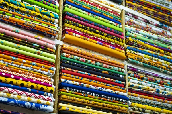 Tekstil pazarındaki renkli desenli kumaş rulolarının yakın çekim görüntüsü. Ekranlar çeşitli renkler, tasarımlar ve dokularla sıralanmış..