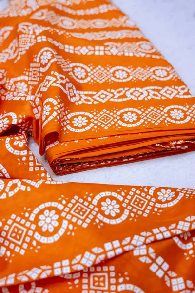 Katlanmış turuncu bir tekstil, büyüleyici beyaz desenli bir tasarım içeriyor. Kumaşın dokusu ve karmaşık ayrıntıları çok güzel sergileniyor, zengin rengi ve zarif tasarımı vurguluyor. Moda için ideal