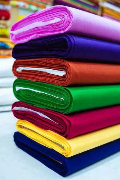 Yakın plan çekimde renkli kumaş rulolarının düzgünce dizilmiş olduğu görülüyor. Her rulo pembe, mor, turuncu, yeşil, kırmızı, sarı ve mavi olmak üzere farklı bir katı renk gösterir. Tekstil pürüzsüz ve sıkı görünüyor.