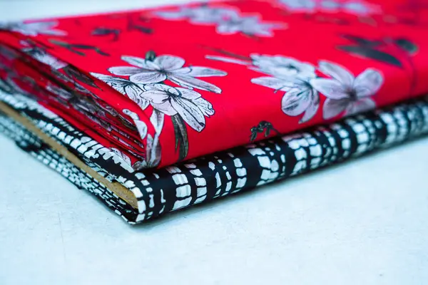 Aydınlık bir yüzeye istiflenmiş iki kumaş rulonun yakın görüntüsü. Üst rulo beyaz ve gri çiçeklerin çiçek desenleriyle süslenmiş canlı kırmızı bir arkaplan içerir. Alt rulo siyah ve beyaz geometrik deseni gösterir