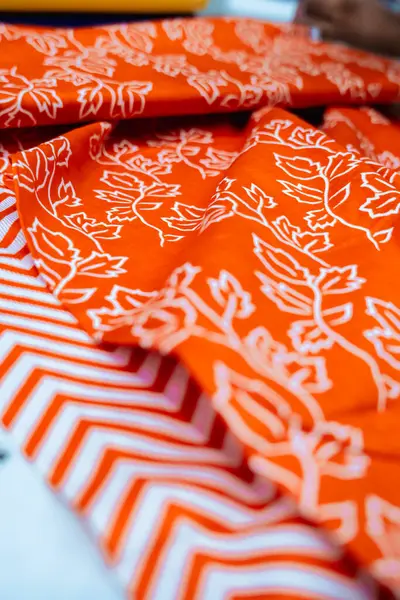 Katlanmış turuncu kumaşın yakın çekim görüntüsü iki farklı deseni gösteriyor. Birinde tekrar eden beyaz çiçekli bir tasarım var, diğerinde ise beyaz ve turuncu renkli kalın bir sembol var. Dokular ve renkler zengindir.