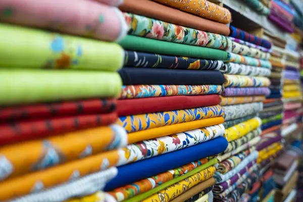 Yakın çekim her biri farklı desenlere ve dokulara sahip renkli kumaş rulolarını ortaya çıkarır. Renklerin ve tasarımların çeşitliliğini yansıtan canlı ve canlı bir ruh hali var. Dekor, düzgün ruloları olan bir tekstil pazarı ya da dükkan gibi görünüyor. 