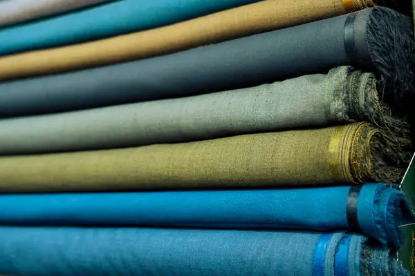 Yakın çekim, düzgün dizilmiş kumaş rulolarını ortaya çıkarır. Her rulo, koyu mavi ve yeşilliklerden sessiz altın ve grilere kadar uzanan eşsiz bir renk ve doku sergiliyor. Kumaşlar sıkıca sarılmış.