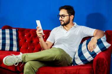 Gözlüklü rahat genç bir adam canlı mavi bir duvara karşı lüks bir kırmızı kadife koltukta rahatça oturur. Sıradan bir şekilde akıllı telefonunu tutar ve bir anlık dijital rahatlık ve memnuniyet göstererek kullanır. Sahne sakin ve davetkar.,