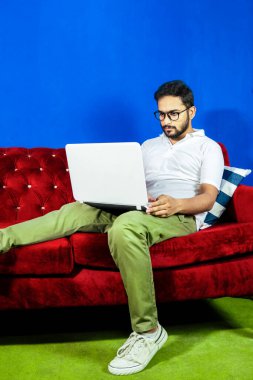 Koyu renk saçlı ve sakallı bir adam, parlak mavi arka planda, kırmızı kadife bir kanepede oturuyor. Gıcır gıcır beyaz bir polo tişört ve zeytin yeşili pantolon giyiyor ve ayakları temiz beyaz spor ayakkabılarla kaplı. Dikkatini topladı. 