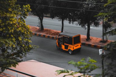An orange auto rickshaw travels down a city street lined with trees. The scene is viewed from an elevated perspective, showing the road, sidewalks, and surrounding vegetation.