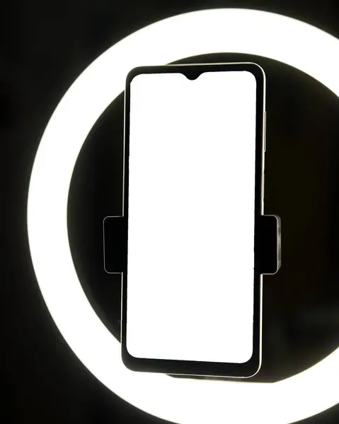 Parlak beyaz ekranlı bir akıllı telefon, dairesel bir halka ışığıyla aydınlatılan koyu bir arkaplan karşısında görüntülenir. Minimalist kurulum uygulama tasarımları, web sitesi arayüzleri veya dijital içerik için idealdir.