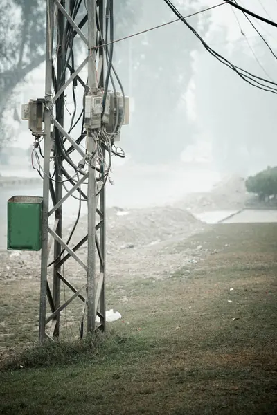 Elektrik kabloları ve alet kutuları ile dolu yıpranmış metal bir elektrik direğinin yakın çekim görüntüsü ön planda belirgin şekilde duruyor. Direğe küçük, koyu yeşil bir kap takıldı. Arka plan puslu, puslu bir şehir manzarası ortaya çıkarıyor. 