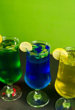 Işıl ışıl mavi, yeşil ve sarı nane kokteylleriyle dolu üç saplı bardak yeşil bir arka planda duruyor. Limon dilimleri her içkiyi süslüyor..