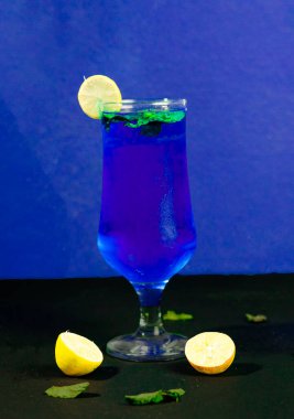 Limon dilimi ve nane filizi ile süslenmiş parlak mavi bir kokteyl ile dolu uzun bir bardağın yakın çekimi. İki limon ve fazladan nane yaprakları koyu mavi bir zemine saçılmış..