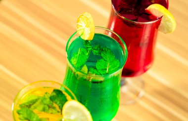 Üç canlı kokteylin yakın plan görüntüsü. Her bardakta farklı renkte bir sıvı var. Taze nane yaprakları ve limon dilimleriyle süslenmiş. İçkiler hafif ahşap bir yüzeye ayarlandı..