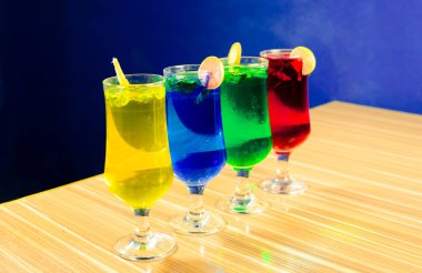 Parlak renkli kokteyllerle dolu dört uzun bardak açık ahşap bir masada koyu mavi bir arka planda oturur. İçecekler sarı, mavi, yeşil ve kırmızı, her biri limon dilimi ve nane ile süslenmiş. Sahne aydınlık.,