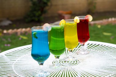 Dört bardak renkli içecek, mavi, yeşil, sarı ve kırmızı, beyaz bir teras masasında düzenlenir. Her içki bir meyve dilimi ve bir tutam nane ile süslenir. Arka plan yemyeşil bir bahçenin bulanık bir görüntüsü..