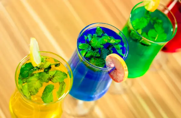 Üç renkli kokteylin yakın plan görüntüsü. Ön planda parlak sarı, mavi ve yeşil sıvılarla dolu üç saplı bardak yer alıyor. Her bardakta taze nane yaprakları ve bir dilim limon ya da benzer turunçgiller var.. 