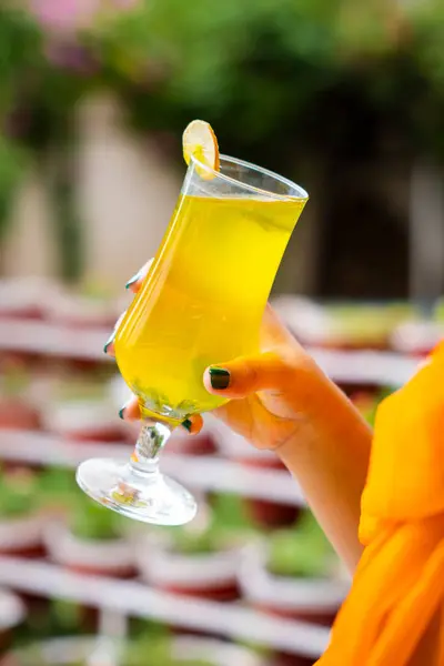 Yakın planda, içi canlı sarı turunçgil içeceğiyle dolu uzun bir bardağı tutan bir el görülüyor. İnce bir dilim limon ya da limon içeceği süslüyor. Arka plan hafifçe bulanık. Terracotta saksılarındaki küçük yeşil bitkileri gösteriyor.