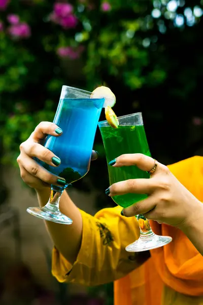 Kokteyl bardaklarını birbirine çarpan iki elin yakın görüntüsü. Bir bardak parlak mavi bir içecek içeriyor, diğeri canlı yeşil bir içecek. Her iki içecek de narenciye ile süslenmiş. Elleri koyu yeşil ojeyle süslenmiş ve sarı elbiseler giymişler..