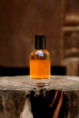 Cam bir kehribar renkli parfüm şişesi yıpranmış bir ağaç kütüğünün üzerinde duruyor. Arka plan bulanık, koyu kahverengi ahşap bir yüzeyi gösteriyor. Şişenin üzerinde ince bir parfüm serpintisi görülebiliyor. Bu da koku ve doğal elega hissini arttırıyor.