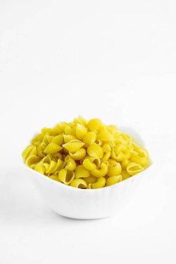 Bir fotoğraf, beyaz arka planda duran, dirsek makarnası olarak da bilinen, pişmemiş gomiti makarnasıyla dolu küçük beyaz bir kase gösteriyor. Makarna parlak sarı, ışık yumuşak ve makarna şeklini ve dokusunu vurguluyor.