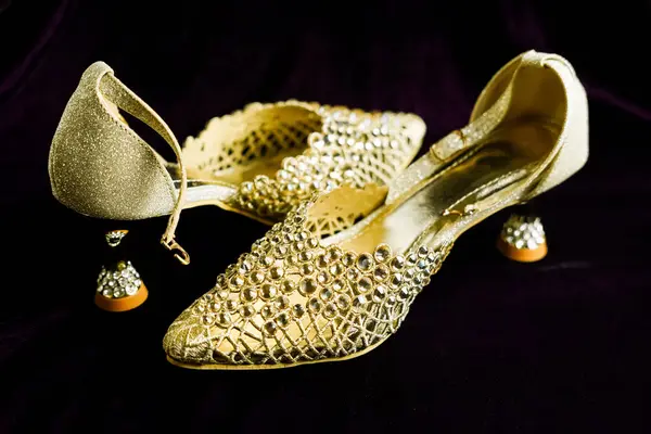 Bir çift zarif kadının topukları koyu bir arka planda sergileniyor. Ayakkabılar altın, parlak kristallerle süslenmiş ve hassas bir ayak bileği kayışı içeriyor. Topuklar düşük ve kristaller stratejik olarak yerleştirilmiş, lüks bir şey yaratıyor.