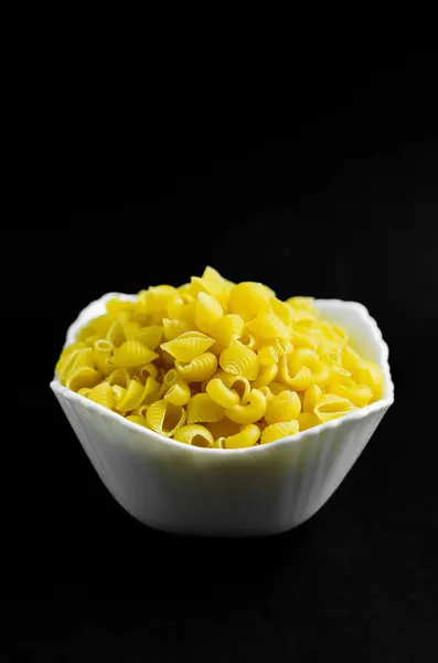 Yakın plan çekimde, pişmemiş makarna kabuklarıyla dolu beyaz, flütlü bir kase ortaya çıkıyor. Canlı sarı renkli makarna, kasenin ortasına cömertçe istiflenmiştir. Mermiler küçük ve hassas, kıvrımlı bir şekil sergiliyorlar. Kase koyu bir yüzeyde duruyor, profesyonel.