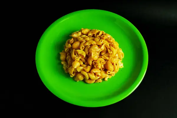  Parlak yeşil bir tabakta duran baharatlı makarna servisinin canlı yakın plan çekimi, katıksız siyah arka planda. Görüntü makarnanın dokusunu ve rengini vurgulayan basit ama çarpıcı bir görüntü sunuyor. Yemek için mükemmel.