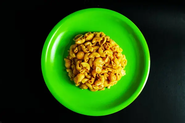 Canlı yeşil bir tabakta pişmiş makarna servisinin yakın çekim görüntüsü. Makarna baharatlı görünüyor ve tabağın ortasında bir tepecikte sunuluyor. Resim, t 'ye güçlü bir karşıtlık sağlayan koyu bir arkaplan üzerine kurulur