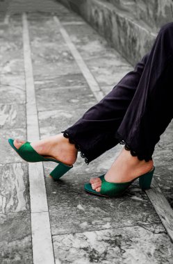 Zümrüt yeşili katırlar ve dantelli siyah pantolonlar giyen birinin bacakları yakından görülüyor. Ayaklar farklı çizgilerle taş bir yüzeye yerleştirilmiştir.