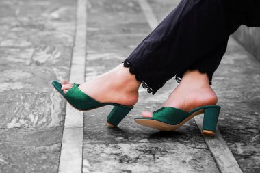 Zarif zümrüt yeşili topuklu sandaletlerle süslenmiş bir kadının ayaklarının yakın görüntüsü. İnce bir parıltıyla parıldayan ayakkabılar, ince dantelli ince detaylarla siyah pantolonunu tamamlıyor. Tek renkli arkaplan, ayakların canlı yeşili vurguluyor.