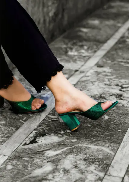  Bir kadının ayaklarının yakın plan görüntüsü şık zümrüt yeşili katırlar ve topuklu ayakkabılar giyiyor. Ayakkabılarda parlak, desenli bir kumaş var. Kadın narin dantelli siyah pantolon giyiyor. Arka planda desenli taş bir yüzey görünüyor