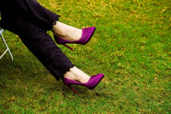   Büyüleyici bir yakın çekim sergisi zarif mor topuklu ayakkabılar ince süslemelerle süslenmiş. Ayakkabılar canlı yeşil bir çimenlikte, görünür şekilde giyinen kişinin bacakları üzerinde, koyu renk pantolonlar ve ince dantelli.