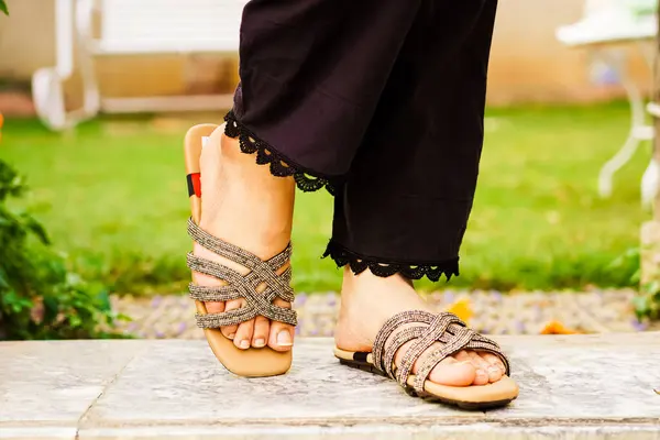 Süslü terlikler giyen bir kadının ayaklarının yakın plan görüntüsü. Sandaletler açık bej renginde, çapraz bağlarla süslenmiş, küçük, parlak boncuklarla süslenmiş. Kadın, altında ince dantelli siyah pantolon giyiyor. Arka taraf.