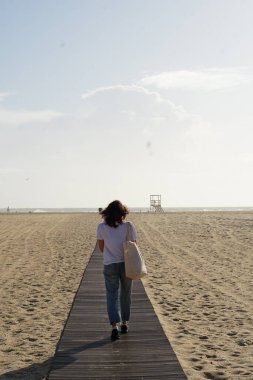 Kumsaldaki esmer kadın, okyanusa giden ahşap yolda yürüyor. 