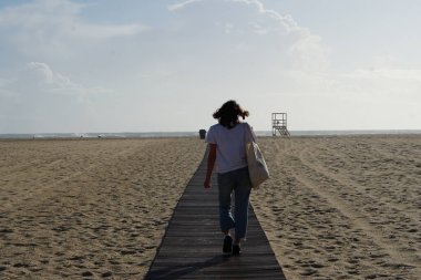 Kumsaldaki esmer kadın, okyanusa giden ahşap yolda yürüyor. 