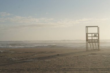 Kumlu bir sahilde ahşap cankurtaran kulesi ve denizin üzerinde günbatımı gökyüzü. 