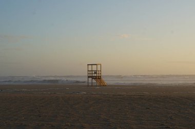 Kumlu bir sahilde ahşap cankurtaran kulesi ve denizin üzerinde günbatımı gökyüzü. 