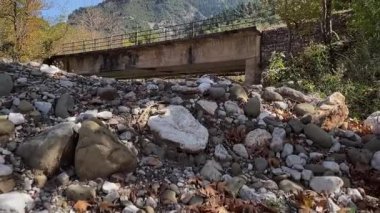 Eski, paslı beton ve çelik bir köprü, arka planda dağ olan ağaçlarla çevrili kurumuş bir nehir yatağının üzerinde duruyor. Doğada unutulmuş bir yapı.