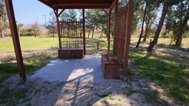 Doğa parkında bankları olan gölgeli bir pergola. Alçak bitki örtüsü, uzun ağaçlar, ve parlak bir bahar ya da yaz günü.