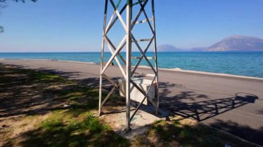 Paslı bir demir deniz feneri ağaçlarla dolu bir sahilde duruyor. Deniz ve yüksek dağlar parlak ve güneşli bir günde çarpıcı bir zemin oluşturur..