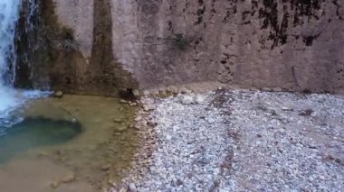 Kayalar, çakıl taşları ve yemyeşil bir vadiyle çevrili geleneksel bir taş duvarın üzerinden akan bir şelale tarafından oluşturulan küçük bir göletin yakın çekimi..