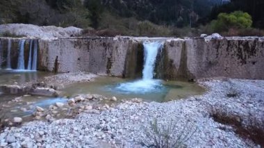 Kayalar ve çalılarla çevrili geleneksel bir taş baraj tarafından oluşturulan küçük şelaleli bir nehrin hava görüntüleri..